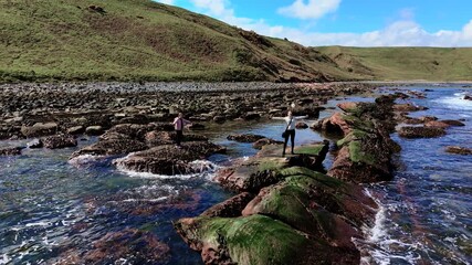 Woman and girl shouting on rocky seashore with arms wide open, releasing stress. Mother and daughter standing by ocean letting go of emotions. Family improving mental health through coastal scream