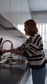 Red-haired woman doing household chores and washing a glass in the kitchen sink with running water
