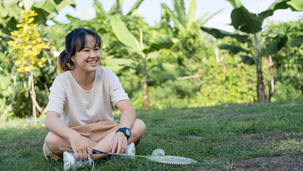 A girl sitting on the grass with a badminton racket, smiling in a natural outdoor garden.