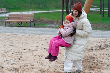 Laughing young mother and daughter spending joyful time together at a playground, playing on a swing, dressed warmly in jackets and hats, enjoying family bonding outdoors, expressing love and support