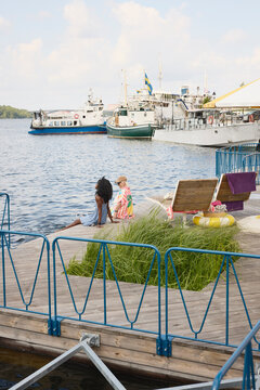 Female friends enjoying leisure time sitting on boardwalk near river with ship in background