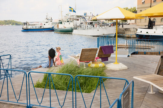 Female friends spending leisure time sitting on boardwalk near river with ship in background