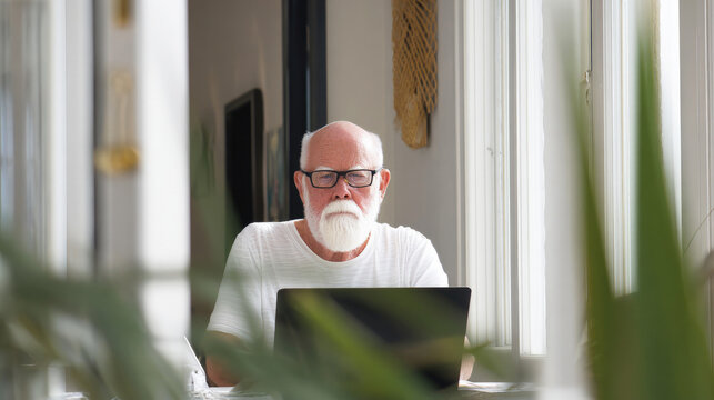 Senior man with white beard working on laptop at home office. Interaction of older people with digital gadgets. - Powered by Adobe