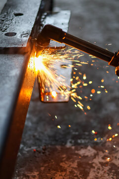 A hand holds a brass cutting torch, generating intense flame and bright sparks while precisely heating and cutting a heavy steel I-beam in a dark workshop.