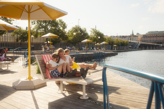 Mother and son spending leisure time with each other while sitting on deck chair near river at sunny day