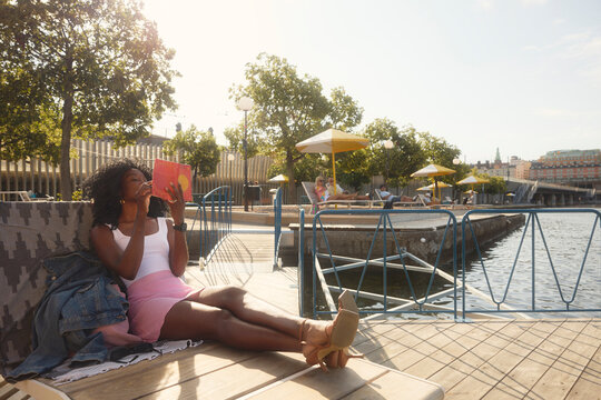 Woman comfortably reading book while sitting on boardwalk near river at sunny day