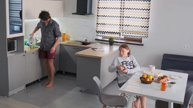 Morning scene in a bright kitchen. A man prepares tea while a woman sits at the table with bread, fruits, and coffee. Peaceful family breakfast, domestic life, cozy lifestyle at home.
