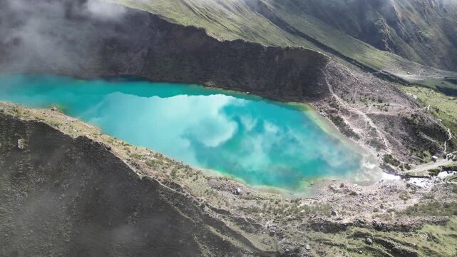 Aerial view of Humantay Lake in the Andes, showing turquoise water reflecting moving cloud patterns beneath sunny skies.