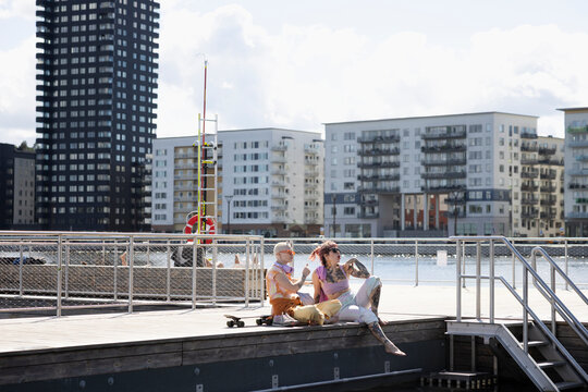 Hipster women spending leisure time with each other while sitting on jetty at sunny day