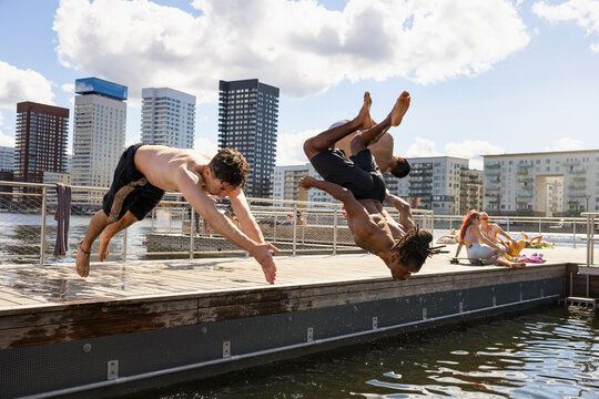 Multiracial male friends diving together in water under cloudy sky