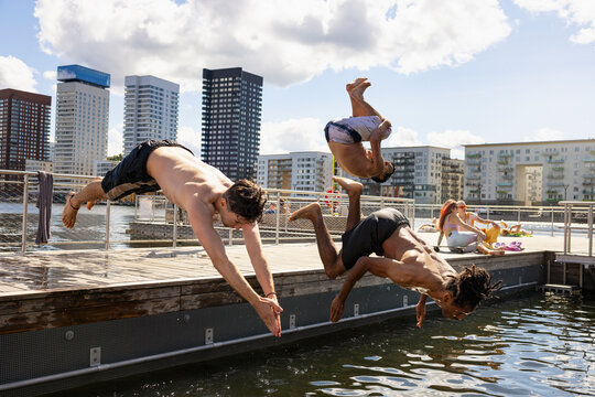 Shirtless male friends diving together in water under cloudy sky at city