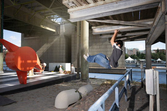 Side view of young man hanging on rod while jumping above railing under bridge