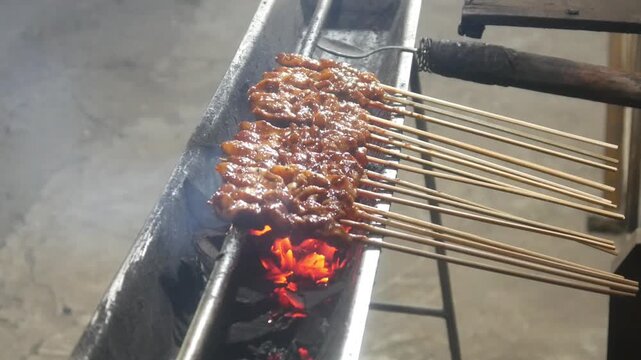 The process of grilling chicken satay over hot coals on a long grill. Rows of skewers are golden brown with a glistening of spices, while flames and smoke rise from the grill.