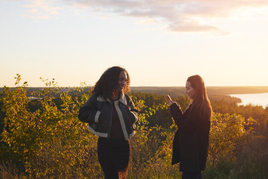 Woman using smart phone standing with friend on hill at sunset