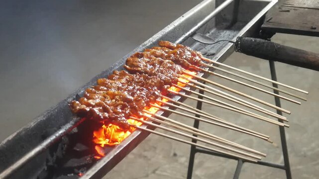 The process of grilling chicken satay over hot coals on a long grill. Rows of skewers are golden brown with a glistening of spices, while flames and smoke rise from the grill.