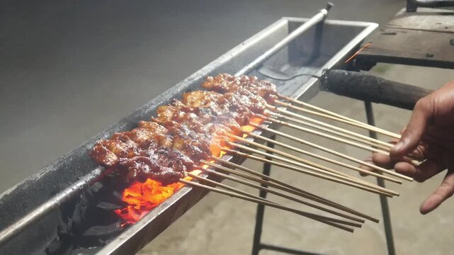The process of grilling chicken satay over hot coals on a long grill. Rows of skewers are golden brown with a glistening of spices, while flames and smoke rise from the grill.