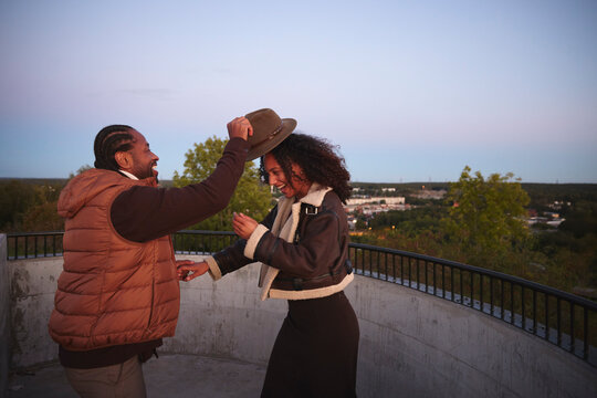 Happy woman putting hat on friend's hat while standing on terrace