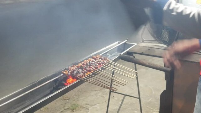 The process of grilling chicken satay over hot coals on a long grill. Rows of skewers are golden brown with a glistening of spices, while flames and smoke rise from the grill.