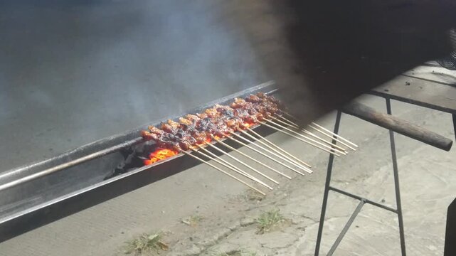 The process of grilling chicken satay over hot coals on a long grill. Rows of skewers are golden brown with a glistening of spices, while flames and smoke rise from the grill.