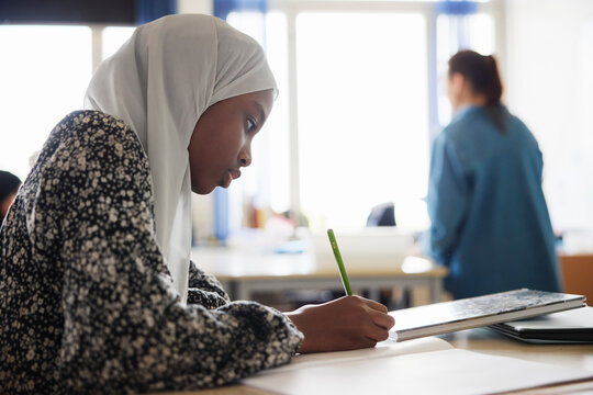 Side view of girl writing in book while sitting in classroom at school
