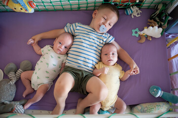 A young boy in a striped shirt and green shorts lies in a crib with his newborn twin brothers. He is holding their hands. Stuffed animals and baby toys surround them