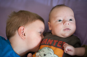 A young boy in a blue shirt cuddles and snuggles his infant baby brother wearing a grey shirt with a character on a purple blanket indoors during the day