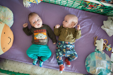 Two cute infants, twins brothers, lie side-by-side in a purple crib, wearing colorful outfits and socks. Stuffed animals and blankets surround them in their sleeping space