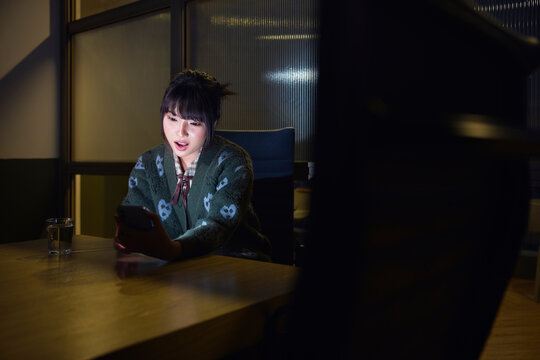 Shocked young businesswoman using smart phone while sitting in dark board room at office
