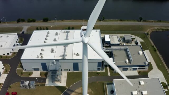 Wind turbine spins above a manufacturing large industrial data center building infrastructure with computing network technology area in a coastal region
