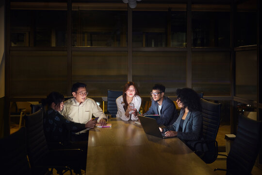 Female business professionals using wireless technologies while sitting in office meeting with male colleagues at night