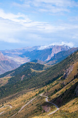 Naklejka premium Picturesque landscape of a dirt road cutting across a steep green mountain slope with rugged peaks and clouds in the distance.