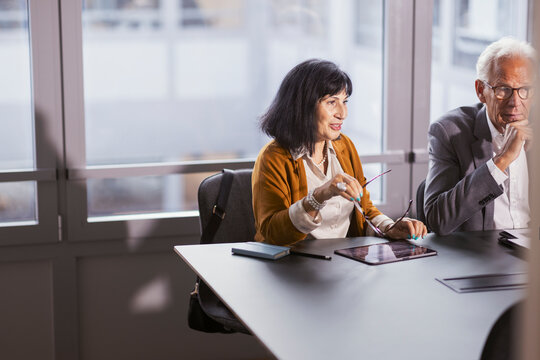 Smiling senior female corporate professional holding eyeglasses talking with colleagues in meeting room at office