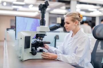 A scientist in a lab coat focuses on the microscope while analyzing samples at a desk. The laboratory has modern equipment and bright lighting.