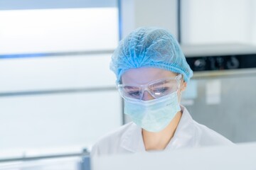 A healthcare worker is focused on a task in a lab, wearing a face mask and protective glasses. The worker is engaged in a biochemistry analysis while dressed in a lab coat and bonnet.