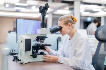 A scientist looks through a microscope at samples on a table in a laboratory. Other researchers work in the background, and the space is well-lit with modern equipment.