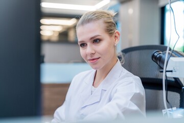 A young woman in a white lab coat sits at a desk using a computer in a laboratory. She appears focused on her work while surrounded by scientific tools.