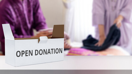 Open a donation box with the words OPEN DONATION printed on the side, with blurred figures in the background sorting clothes for a charity drive. Concept of Open Donation