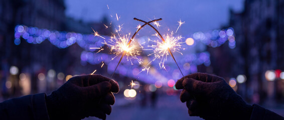 Hands holding festive sparklers at dusk with blurred city street lights and blue holiday decorations creating a magical celebration mood