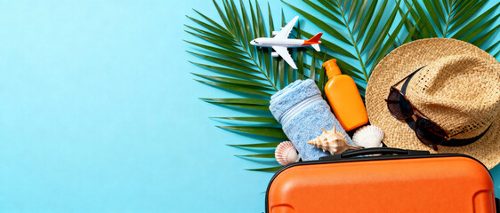 Summer travel essentials with orange suitcase, tropical palm leaves, sunscreen, hat, towel, sunglasses, and miniature plane