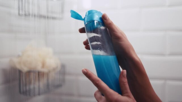 Close-up of a hand opening a blue shampoo bottle in the shower, revealing the product. Self-care concept and refreshing bathroom atmosphere.
