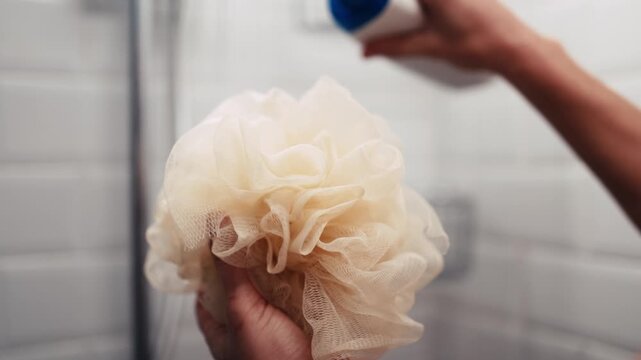 Person holds a soft loofah while applying shower gel from a bottle, highlighting a refreshing bathing routine. Self-care and bath cosmetics advertising concept.