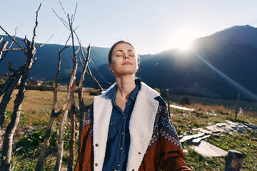 Naklejka premium Woman standing in mountains with sunlit outdoors scene, enjoying nature in a meadow wearing a warm jacket, eyes closed in relaxation and peaceful outdoor portrait at golden hour