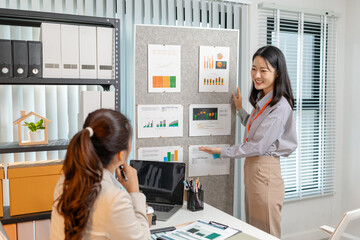 Two Asian businesswomen are smiling while discussing company performance reports in a modern office. They analyze data from printed documents, showing teamwork, planning, and collaboration in business