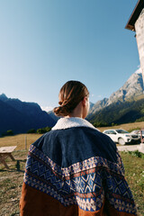 Naklejka premium Woman back looking at mountains in the countryside, wearing a patterned jacket. Outdoors travel scene with parked car, alpine valley, rural house and clear summer sky.