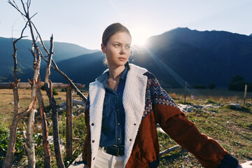 Naklejka premium Woman portrait in countryside with mountains and sunlight, wearing a shearling jacket and denim shirt by a rustic fence. Outdoor fashion, thoughtful expression and natural scenery.