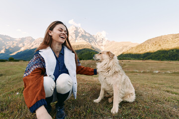 Naklejka premium woman dog mountains nature smiling petting meadow outdoors: young woman kneeling in alpine meadow laughing as she pets her dog with scenic mountain range and clear sky behind