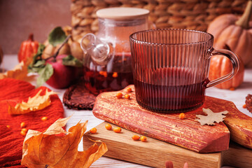 Ribbed glass mug with red herbal tea and teapot in background on rustic wooden surface with autumn leaves and apple
