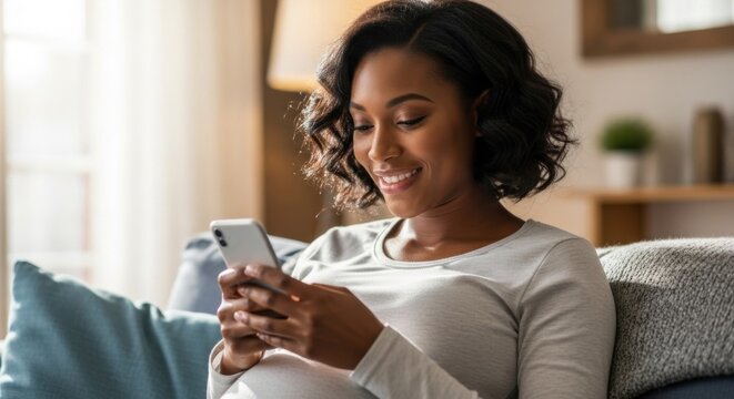 Happy pregnant african american woman using smartphone sitting on sofa at home. Expecting mother texting or checking app on mobile phone. Maternity and modern technology concept. - Powered by Adobe