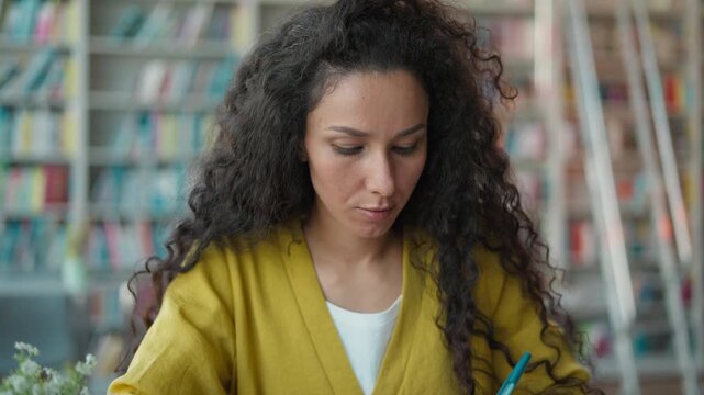 Focused Indian Hispanic woman writing notes pen in library studying female girl student learning homework notebook textbooks knowledge research high school university bookstore writer focus education
