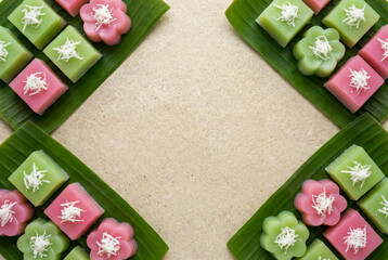 Traditional Thai dessert flat lay of pink and green Khanom Chan layer cakes on banana leaves with shredded coconut topping and copy space in center on beige stone background for food menu design.